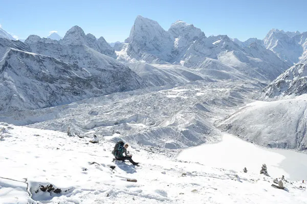 A trekker resting on a snow-covered mountain slope above a vast glacier and frozen alpine lake surrounded by snow-capped peaks