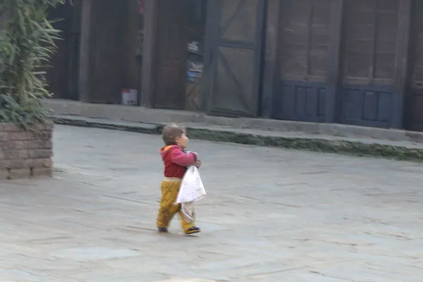 A young child wearing a red jacket and yellow pants walking alone along a stone-paved street in a Nepali village.