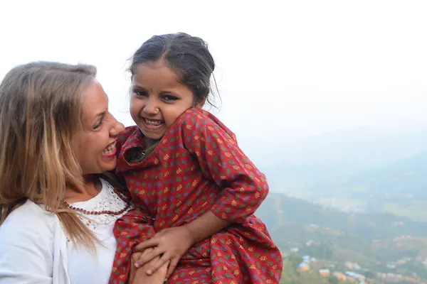 Foreign female traveler laughing with a local Nepalese child in a red patterned dress during a trek in Nepal.