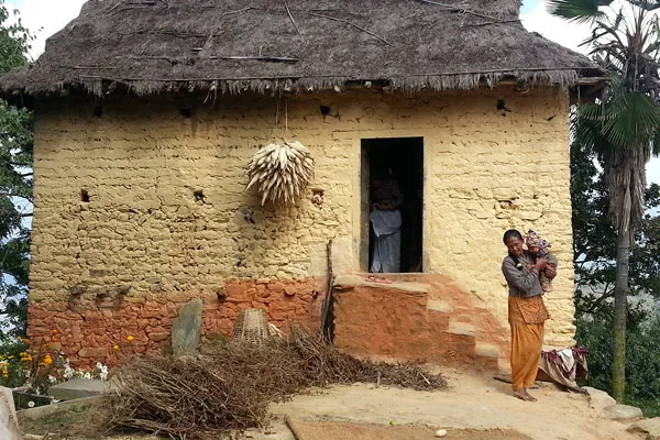 A local Nepalese mother holding her child outside a traditional thatched-roof mud house in an indigenous village