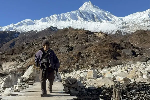 A local man walking across a small wooden footbridge in a rocky Himalayan landscape with a massive snow-capped peak behind him