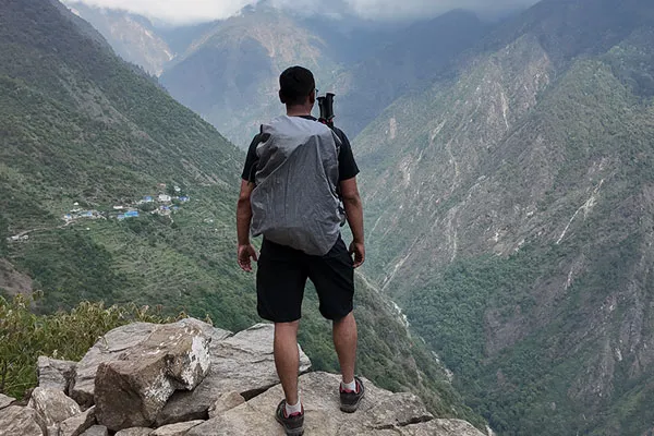 A trekker with a grey backpack standing on a cliff edge looking out over a steep mountain valley in the Langtang region