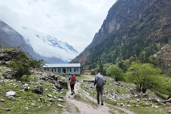 Two trekkers with backpacks and hiking poles walking along a dirt trail toward a teahouse with high green mountain walls and snow-capped peaks behind