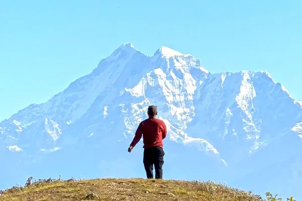 A lone person in a red sweater standing on a grassy hill facing a massive snow-covered mountain peak