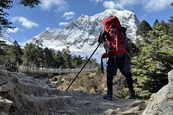 A trekker with a large red backpack and hiking poles standing on a stony trail overlooking a suspension bridge and snow-capped Himalayan mountains