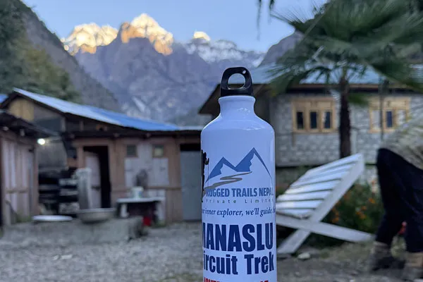 A white branded water bottle labeled Manaslu Circuit Trek standing on an outdoor table in front of a mountain lodge with snow-capped peaks behind.