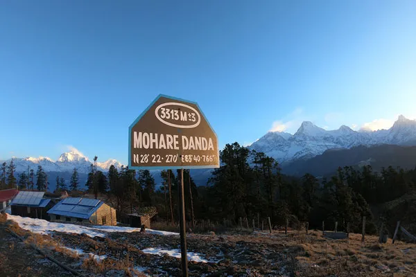 A metal signpost marking Mohare Danda at an elevation of 3313 meters with snow-capped Dhaulagiri and Annapurna mountain peaks under a clear blue sky in Nepal.