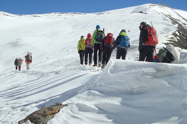 A line of mountaineers carrying heavy backpacks trekking single-file across a high-altitude snow slope