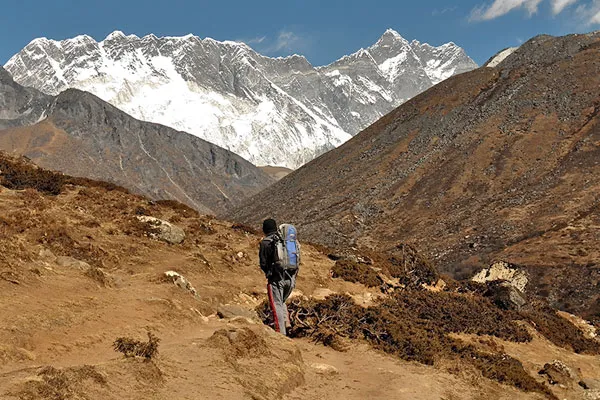 A solitary trekker carrying a backpack walking along a rocky high-altitude path in the Khumbu region toward snow-covered Himalayan peaks.