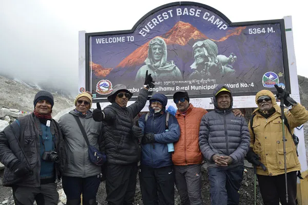 A group of seven male trekkers in heavy winter gear posing in front of the Everest Base Camp welcome sign and marker at 5,364 meters