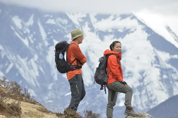 Two hikers in matching orange jackets and backpacks taking a break on a high mountain trail in Nepal with massive snow-covered peaks behind them.