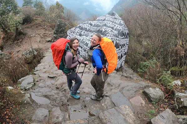Two smiling female trekkers wearing backpacks with bright rain covers next to a Buddhist mani stone in Nepal.