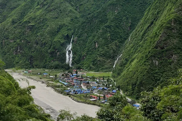 A lush green mountain valley in Nepal with tall waterfalls cascading down steep hillsides past a riverside village during the monsoon season