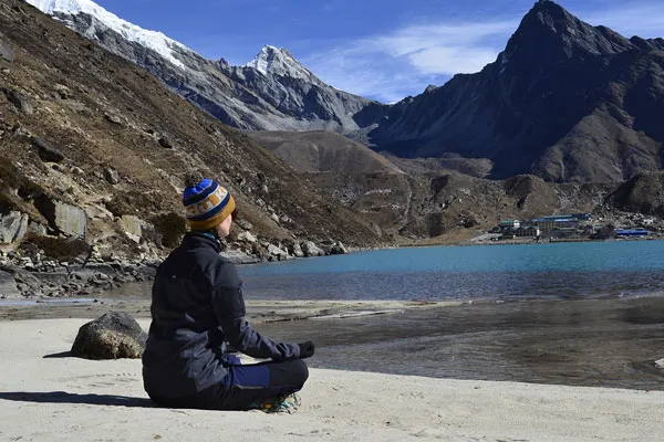 Solo female trekker meditating in a cross-legged position on the sandy shore of Gokyo Lake with snow-capped mountains in the background