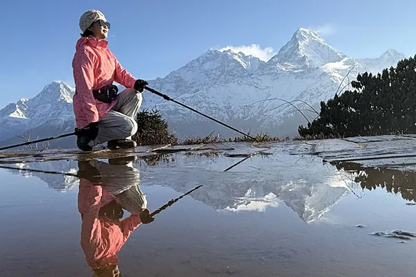 A female trekker in a pink jacket kneeling beside a water puddle that reflects her image and the snow-capped Annapurna mountain range behind her.