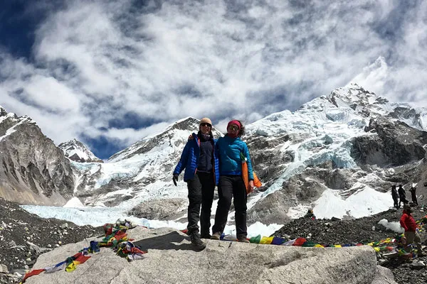 Two women in puffer jackets smiling while standing together on a large rock at Everest Base Camp with Khumbu Glacier in the background