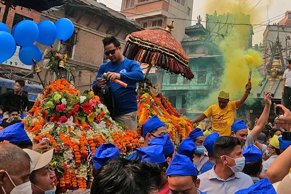 A crowded festival scene during Pahan Chare showing men in matching blue caps carrying a flower-decorated deity palanquin amid yellow smoke and balloons