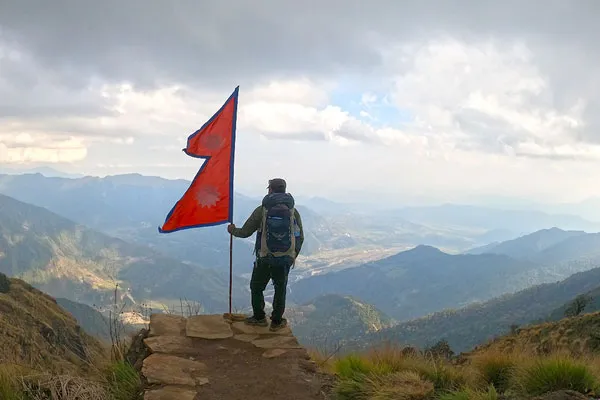 Trekker standing on a rocky cliff edge holding the double-pennant Nepali flag overlooking vast mountain valleys.