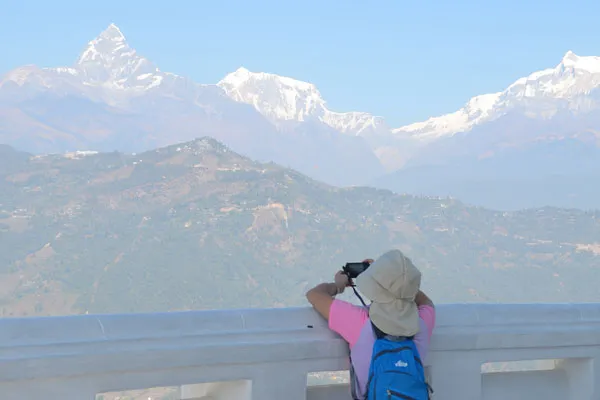 A traveler wearing a sun hat and blue backpack taking a photo of Machhapuchhre (Fishtail Mountain) from a white terrace viewpoint in Pokhara.