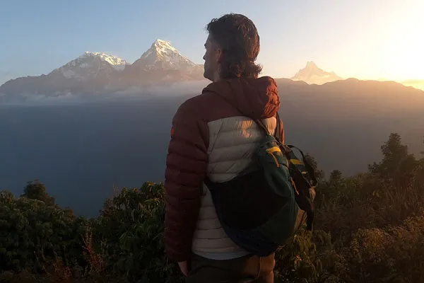 A male hiker wearing a warm padded jacket and backpack standing at a viewpoint observing snow-capped Himalayan peaks