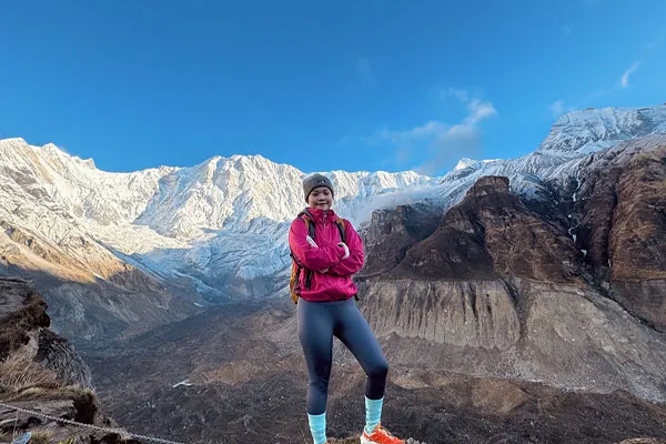 A female trekker in a pink jacket standing on a mountain ridge with massive snow-capped peaks of the Annapurna massif behind her