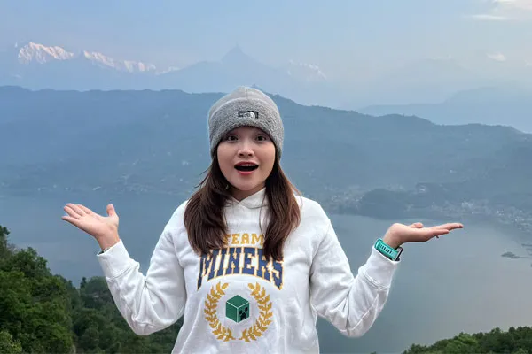 A cheerful young woman in a beanie and white sweatshirt posing with open hands overlooking Phewa Lake and the mountains in Pokhara.