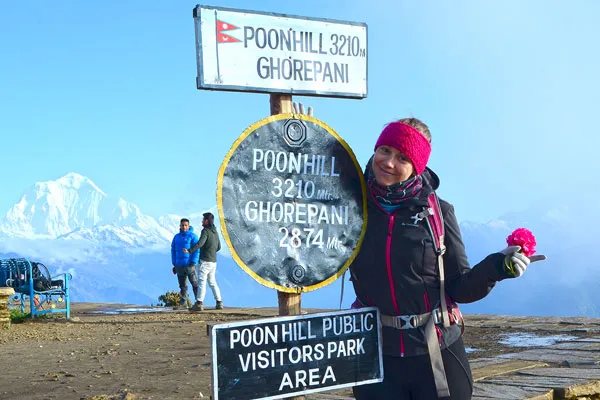 A smiling female hiker posing next to the official Poon Hill 3210m elevation sign with snow-covered Himalayan peaks in the background.