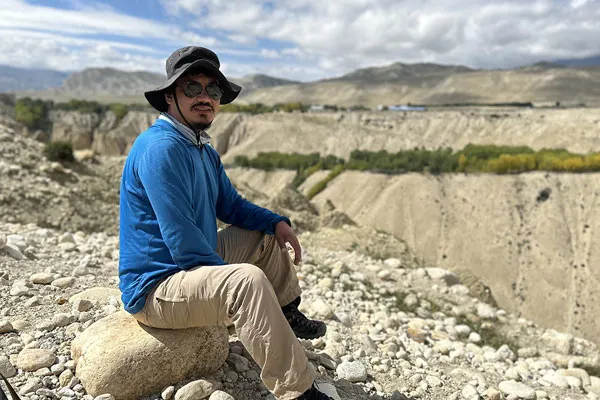 A male guide wearing a sun hat and blue long-sleeve shirt sitting on a rock in the arid desert terrain of Upper Mustang