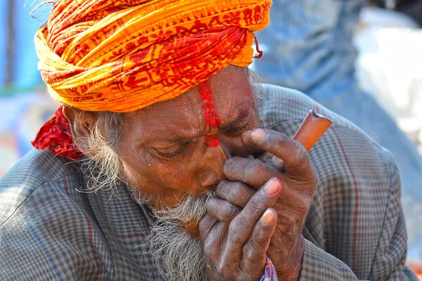 Elderly Sadhu with a saffron turban and red tilak smoking marijuana from a traditional chillum pipe during Maha Shivaratri in Nepal.