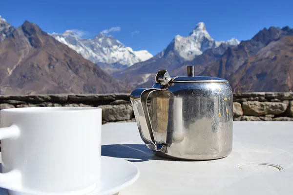 A stainless steel teapot and white coffee mug on a outdoor table facing snow-capped mountain peaks including Ama Dablam under a clear blue sky