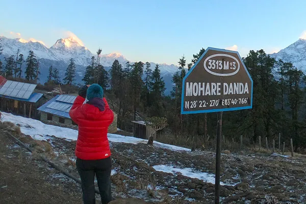 A trekker in a red jacket photographing the snow-capped Annapurna mountain range next to a brown Mohare Danda signpost showing 3,313 meters elevation