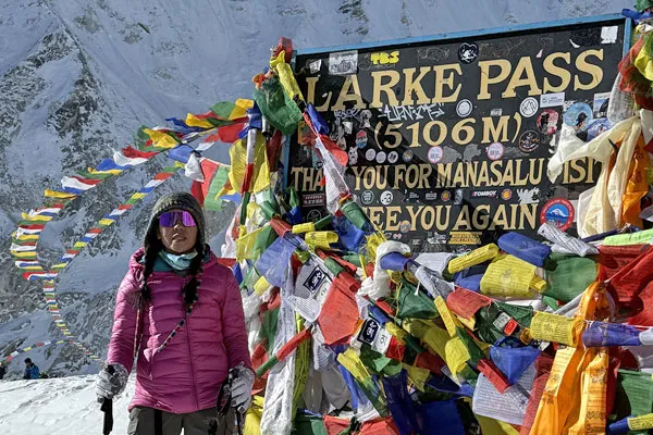 A female trekker wearing a pink jacket and sunglasses posing proudly next to the prayer-flag covered Larkya Pass sign board.