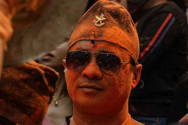 Close-up portrait of a man wearing sunglasses and a traditional Dhaka topi hat, his face covered in bright orange sindoor powder during the Sindoor Jatra festival in Thimi