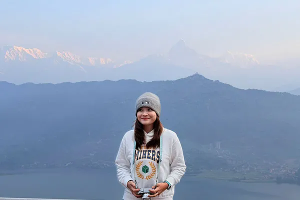 Young woman wearing a beanie holding a camera standing at Sarangkot viewpoint with Machhapuchhre in the background.
