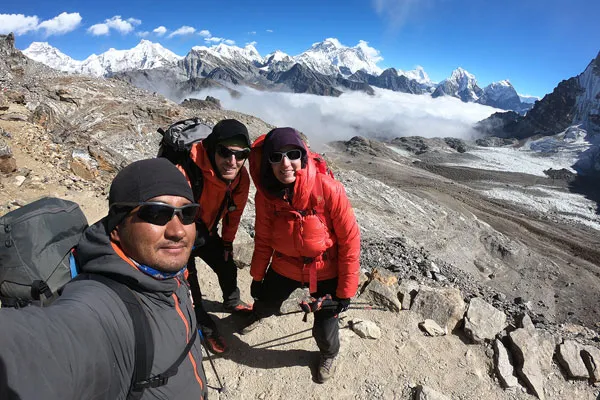 A local Nepali guide taking a group selfie with two foreign trekkers in red down jackets on a high mountain pass overlooking a glacial valley