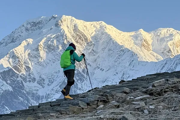 A solo hiker with green backpack and trekking poles climbing stone trail stairs with massive snow-capped mountains illuminated by sunlight ahead