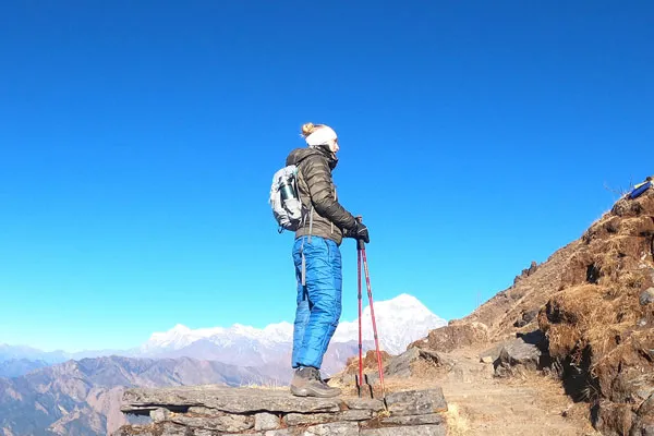 A female trekker in blue insulated pants and a jacket standing on a stone platform holding trekking poles while looking out at distant Himalayan peaks