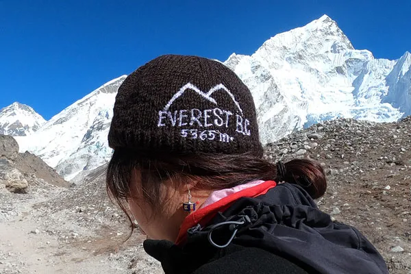 Close-up profile of a trekker wearing a dark brown beanie embroidered with "Everest BC 5364m" against snow-capped peaks