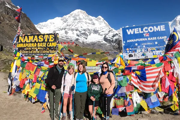 A family of five with trekking poles posing in front of the Annapurna Base Camp welcome signboard covered in colorful prayer flags and international flags