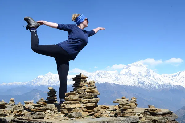 A woman balancing in a yoga dancer pose on a rocky high peak beside stone cairns with the snow-capped Annapurna mountain range