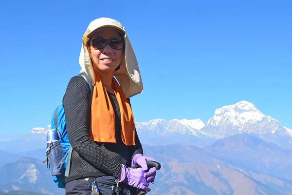 Female trekker with sun protection hat, sunglasses, and trekking poles posing against the Dhaulagiri and Annapurna mountain ranges