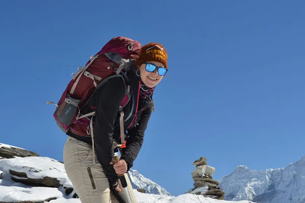 Female trekker with a large backpack, beanie, and sunglasses posing cheerfully on a snowy Himalayan pass.