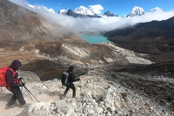 Two trekkers with backpacks and trekking poles hiking down a steep rocky path toward a turquoise glacial lake in Nepal.