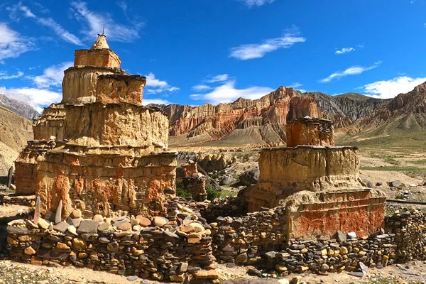A multi-layered ancient clay chorten built above a stacked stone wall set against dramatic red rock cliffs and a bright blue sky in Upper Mustang, Nepal