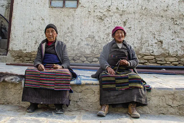 Two elderly Mustang women in traditional Tibetan aprons sitting against a mud-brick house holding prayer beads.