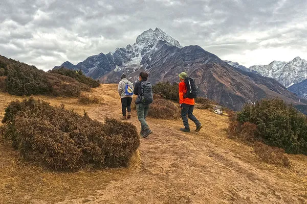 Three trekkers wearing daypacks walking along a dirt mountain trail with a prominent snow-capped peak in the background