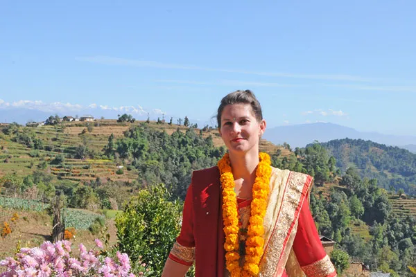 A female traveler wearing a traditional red Nepalese sari and yellow marigold garland posing in front of green terraced hills