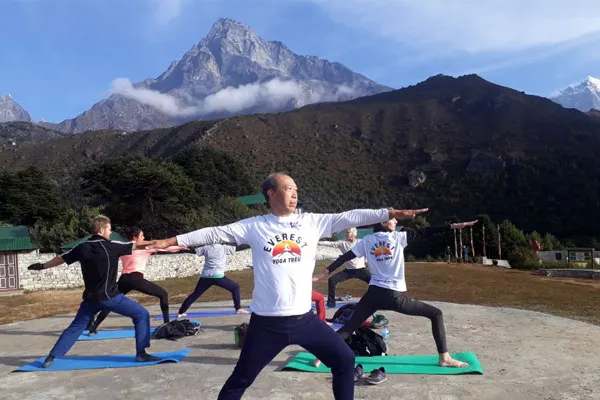 A group of yogis wearing white Everest Yoga Trek shirts practicing Warrior II pose on mats on a stone platform with Himalayan mountains behind them