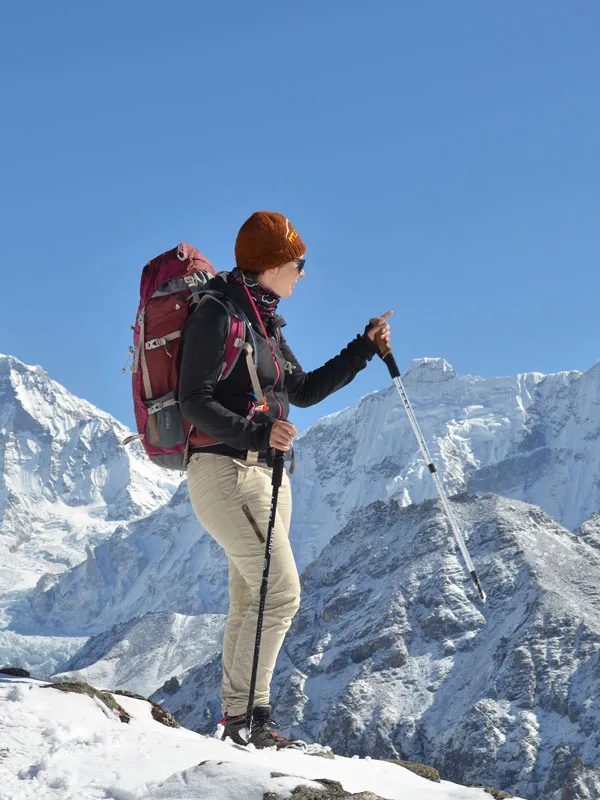 Female trekker with backpack and trekking poles standing on a snowy mountain ridge under a clear blue sky