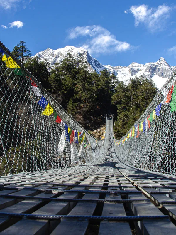 Low-angle perspective looking down a long metal suspension bridge draped with prayer flags leading toward snow-capped mountains